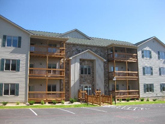 A multi-story building is presented, featuring a combination of wooden balconies and stone accents. Multiple windows allow for natural light, while landscaped greenery surrounds the entrance, creating an inviting exterior. A central entrance is flanked by wooden railings leading to a pedestrian pathway.