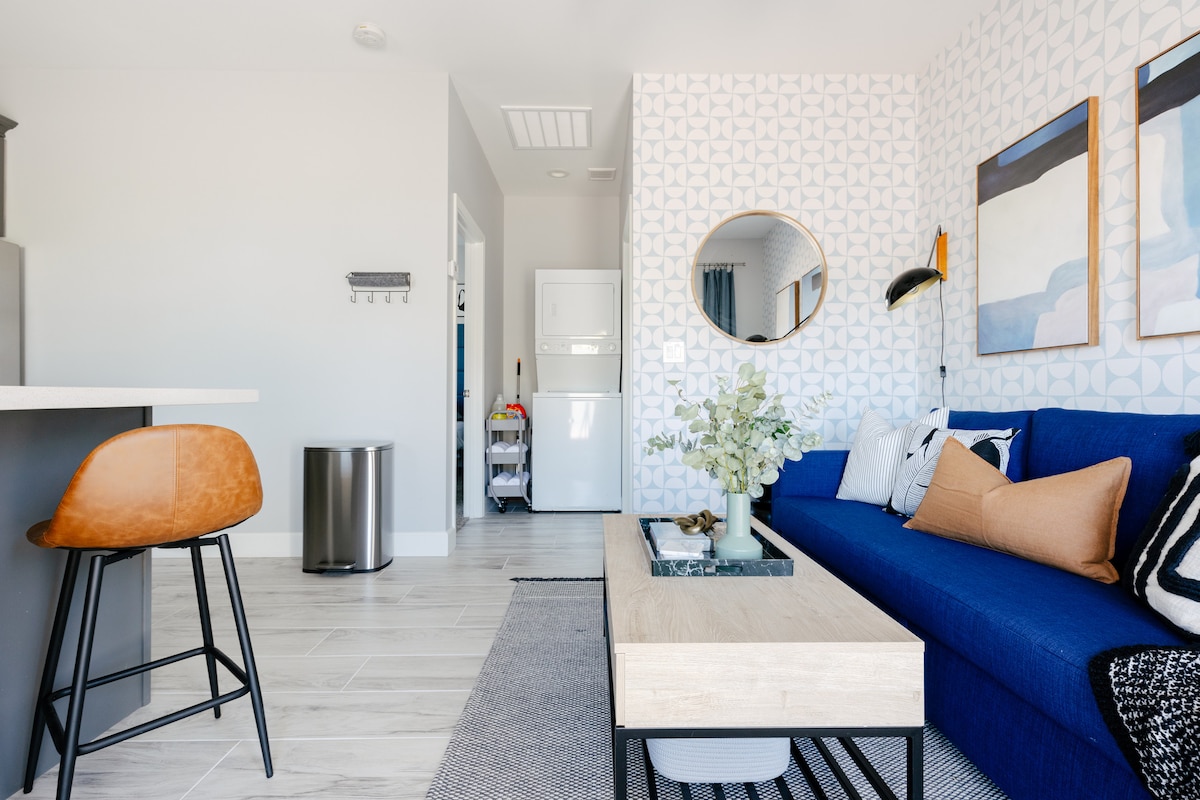 A modern living area features a deep blue sofa accented with neutral throw pillows. A round mirror reflects light, while a geometric wall design adds visual interest. A wooden coffee table sits atop a textured rug, complemented by a stylish bar stool nearby.
