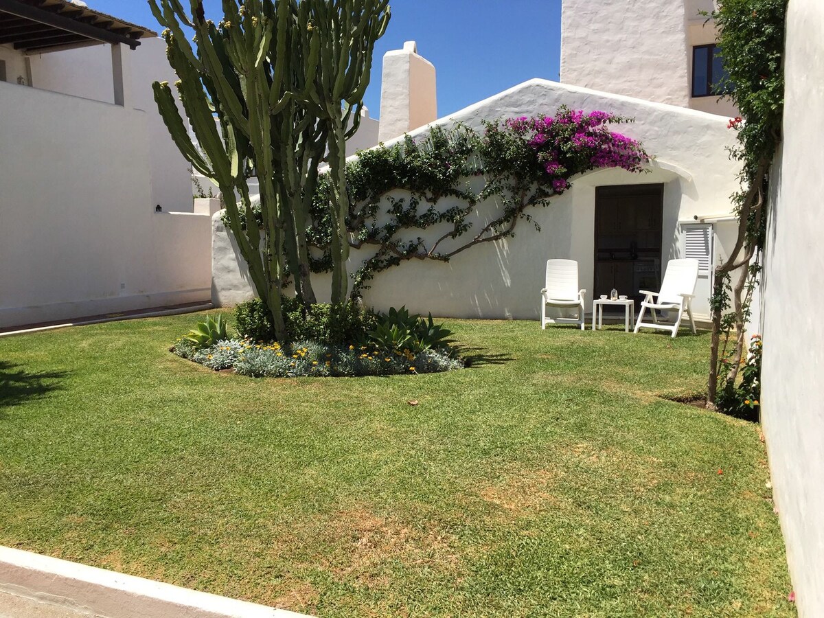 A well-maintained garden area features manicured grass and vibrant flowering plants. Two white chairs are positioned on a small patio, offering a cozy spot for relaxation. A tall cactus and a climbing plant enhance the outdoor space against a backdrop of white walls.