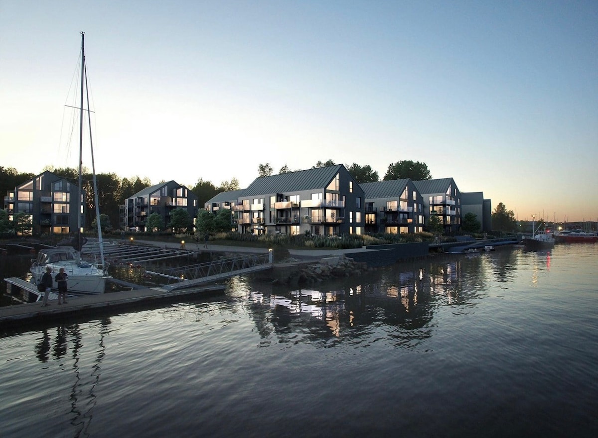 A modern waterfront property is showcased along the riverbank. Several building units feature large windows reflecting the soft evening light. A sailboat is docked nearby, with a wooden walkway connecting the structures. The serene water reflects the architectural lines and trees lining the background.