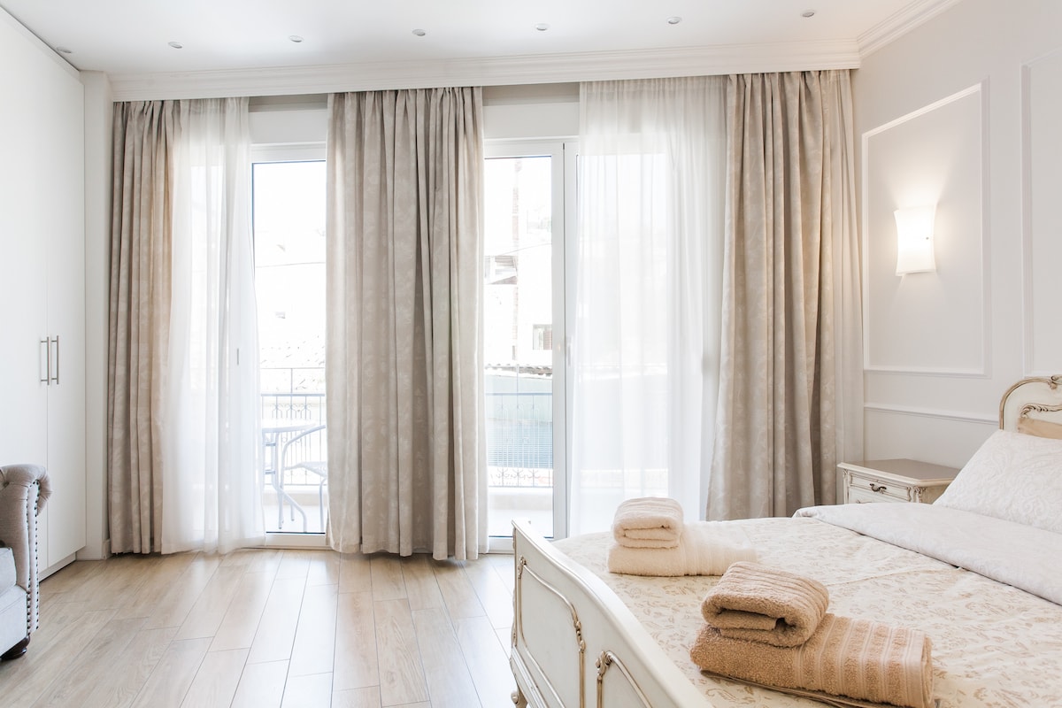 A well-lit bedroom features large windows, allowing natural light to illuminate the space. Soft, neutral curtains frame the windows, complementing the light-colored walls. A spacious bed is dressed in linens, with neatly folded towels placed on its surface. A subtle elegance is showcased in the room's design.