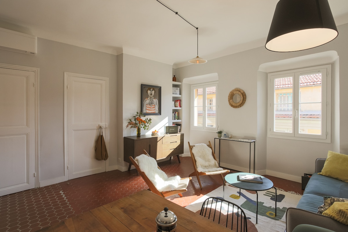 A bright and airy living space features large windows that invite natural light. A comfortable sofa is complemented by two cozy chairs, along with a wooden dining table. Wall art and decorative elements add character to the room, creating an inviting atmosphere.