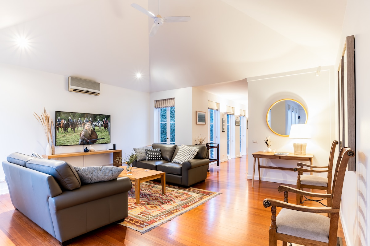 A spacious living area is presented, featuring two comfortable sofas and stylish armchairs positioned around a coffee table. A large TV displays a nature program, contributing to the inviting ambiance. Natural light fills the room, highlighting the polished wooden floor and modern decor.