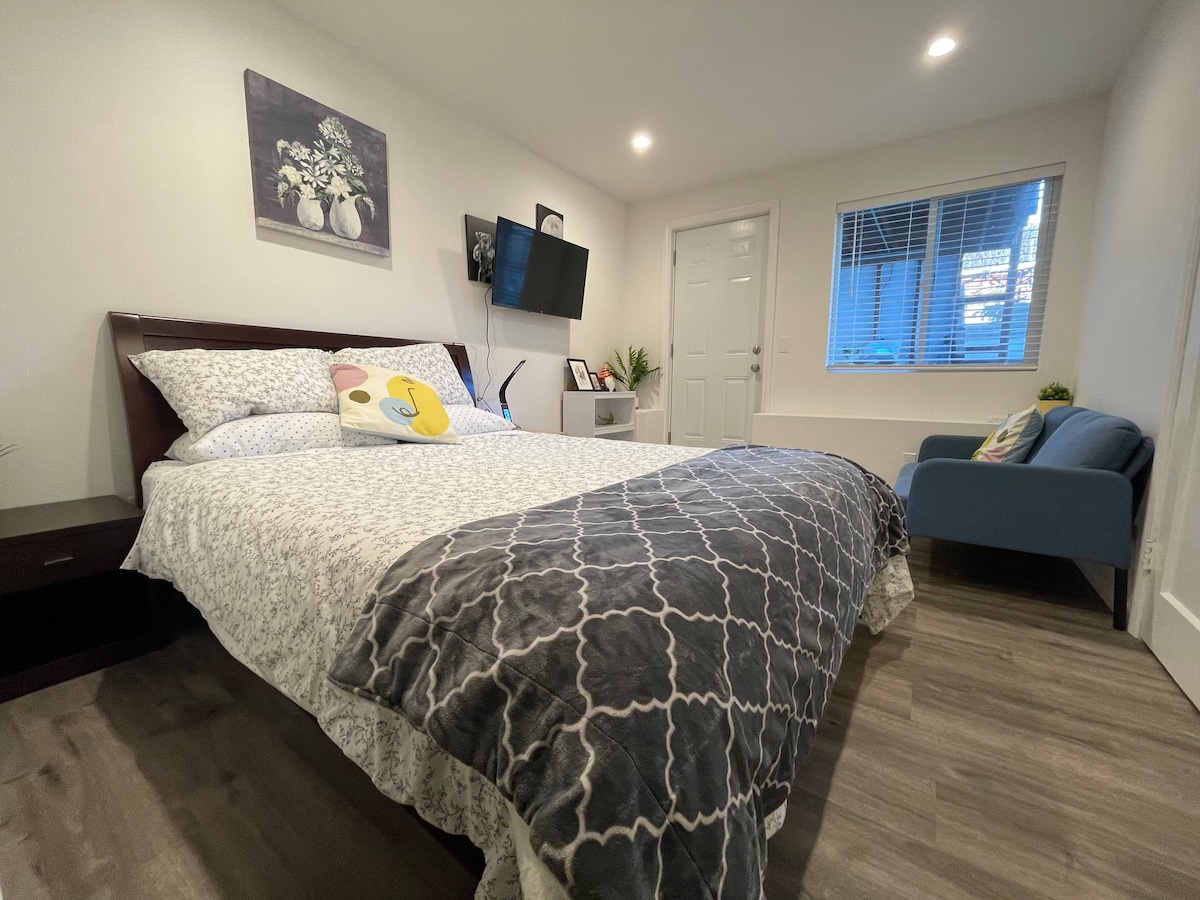 A cozy bedroom is highlighted by a queen-sized bed dressed in patterned linens. A small blue chair is placed near the window, where natural light softly illuminates the room. A television is mounted on the wall opposite the bed, complemented by light-colored walls and modern decor.