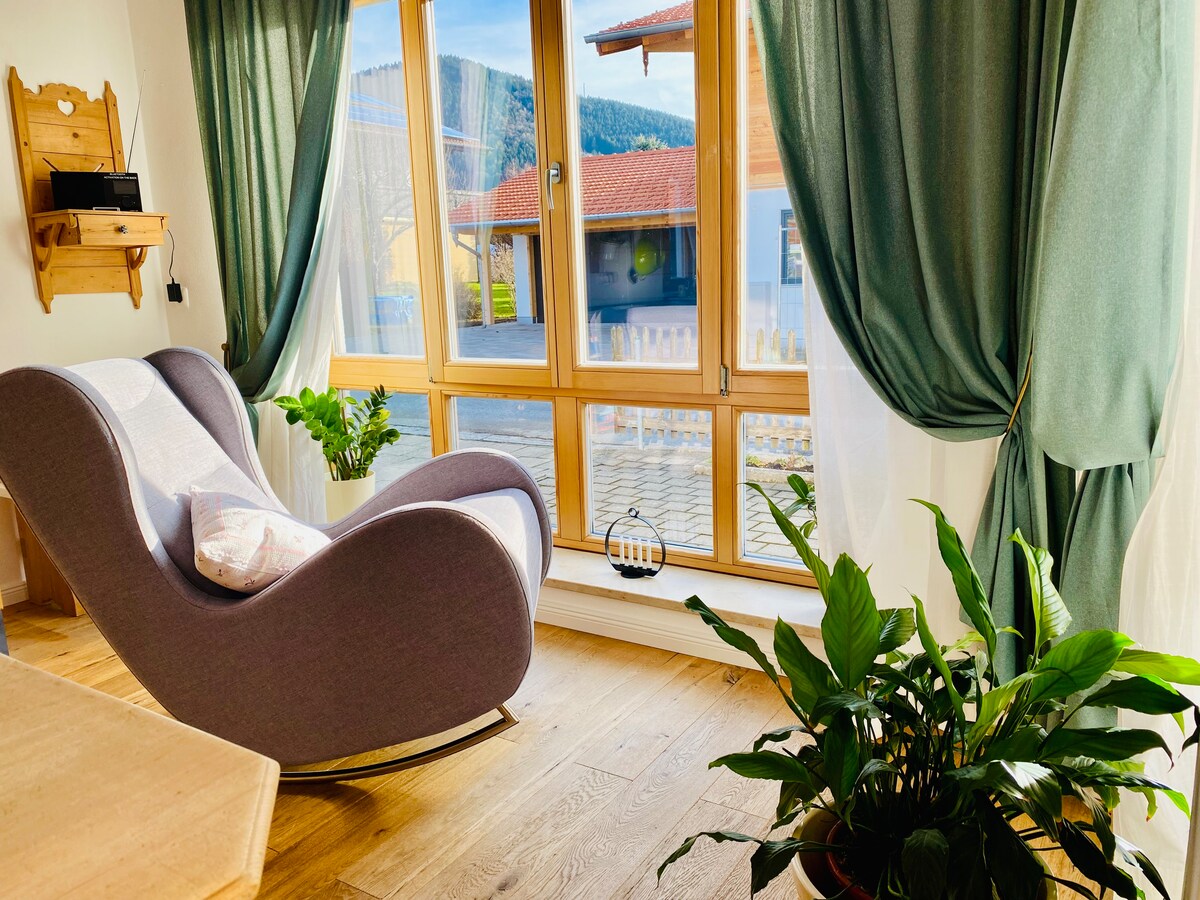A cozy seating area features a modern gray rocking chair positioned beside large wooden windows. The sunlight illuminates the space, highlighting green curtains that frame the view of the outdoors. Potted plants add a touch of greenery to the inviting atmosphere.