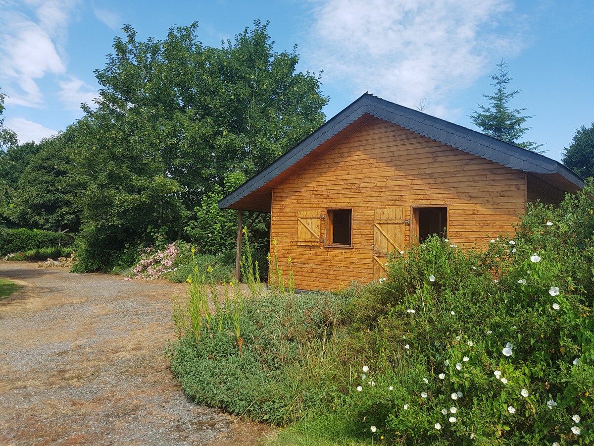 A wooden chalet is set amidst a landscaped area, surrounded by greenery and colorful flowers. The structure features large wooden shutters and a sloped roof. The driveway leads up to the entrance, providing a serene, natural environment.