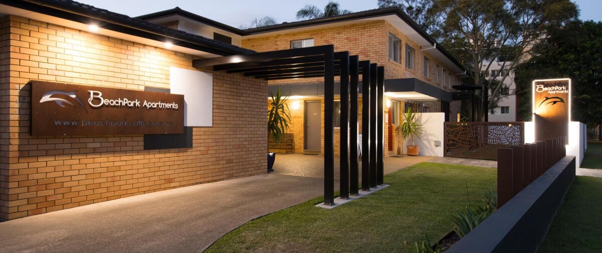 The exterior of BeachPark Apartments is shown, featuring a modern brick facade. An illuminated sign is positioned beside a sleek entrance adorned with vertical beams. Lush greenery adds a touch of nature, while the pathway is lined with well-maintained landscaping, welcoming guests to the property.
