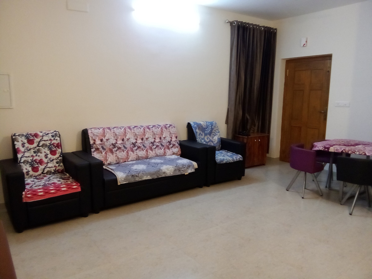 A spacious living area includes two black sofas adorned with floral covers. A small round dining table with purple chairs sits opposite the seating. Natural light is present from a nearby window, and dark curtains frame the space.