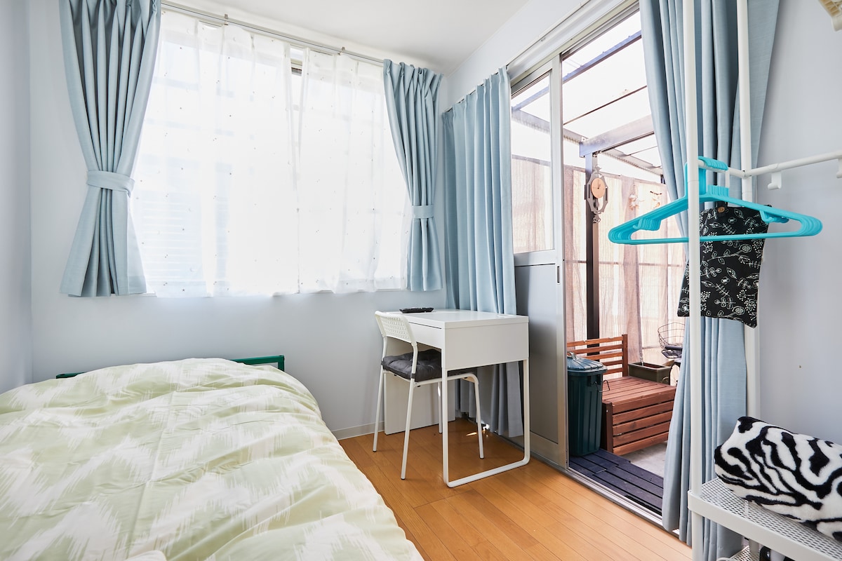 A bright room features a spacious bed with a light green quilt, complemented by sheer curtains. A small white desk and two chairs are positioned near a large window that opens to a wooden terrace, enhancing the indoor-outdoor connection.