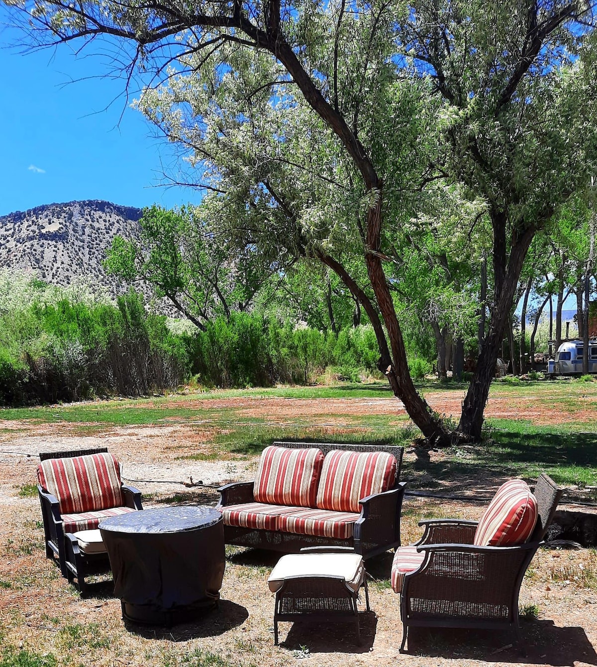 A seating area is set outdoors under a tree, featuring a striped sofa, two armchairs, and a round table. The natural landscape includes green shrubs and a distant mountain. Soft blue sky and scattered clouds enhance the inviting atmosphere.