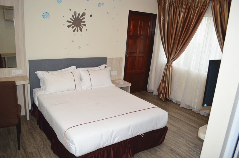 A cozy room features carpeted flooring and wooden furnishings. A spacious bed with white linens is positioned against a light-colored wall adorned with simple decorative details. Natural light filters through sheer curtains, complementing a wooden door and a flat-screen TV.