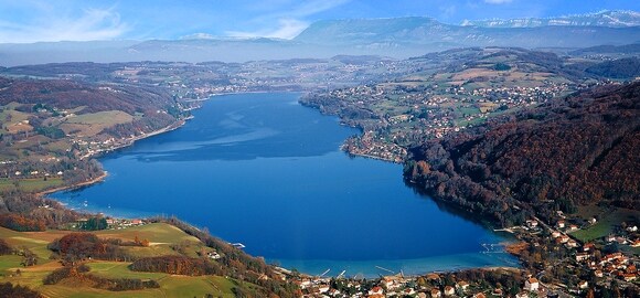 A panoramic view showcases the expansive Lac de Paladru, surrounded by lush green hills and gently rolling landscapes. The tranquil blue water reflects the sky, while charming homes are visible along the shore, contributing to the scenic beauty of the area.