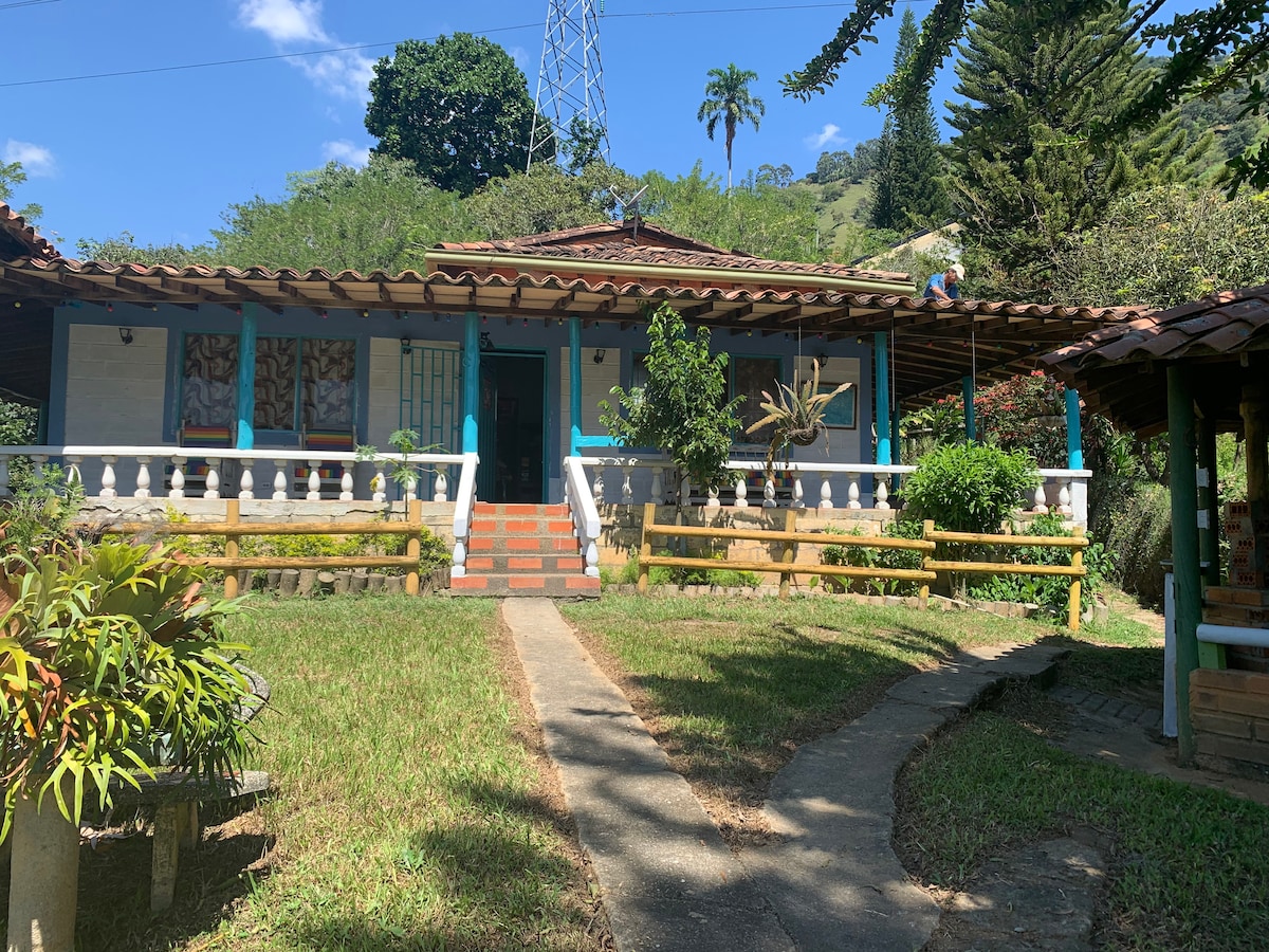 A rustic house is set amid lush greenery, featuring a tiled roof and colorful accents. A pathway leads to the entrance, flanked by decorative plants. The spacious porch offers seating areas and a welcoming atmosphere. Towering trees and distant hills can be seen in the background.