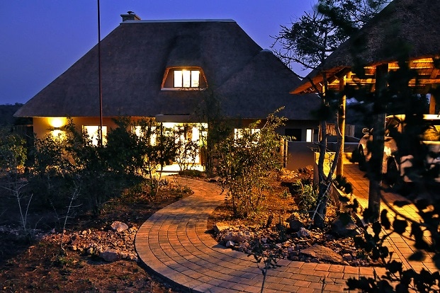 A charming residence featuring a thatched roof is illuminated by soft lighting at dusk. A winding pathway leads to the entrance, bordered by landscaped greenery. Windows reflect the warm glow from inside, suggesting a welcoming ambiance.