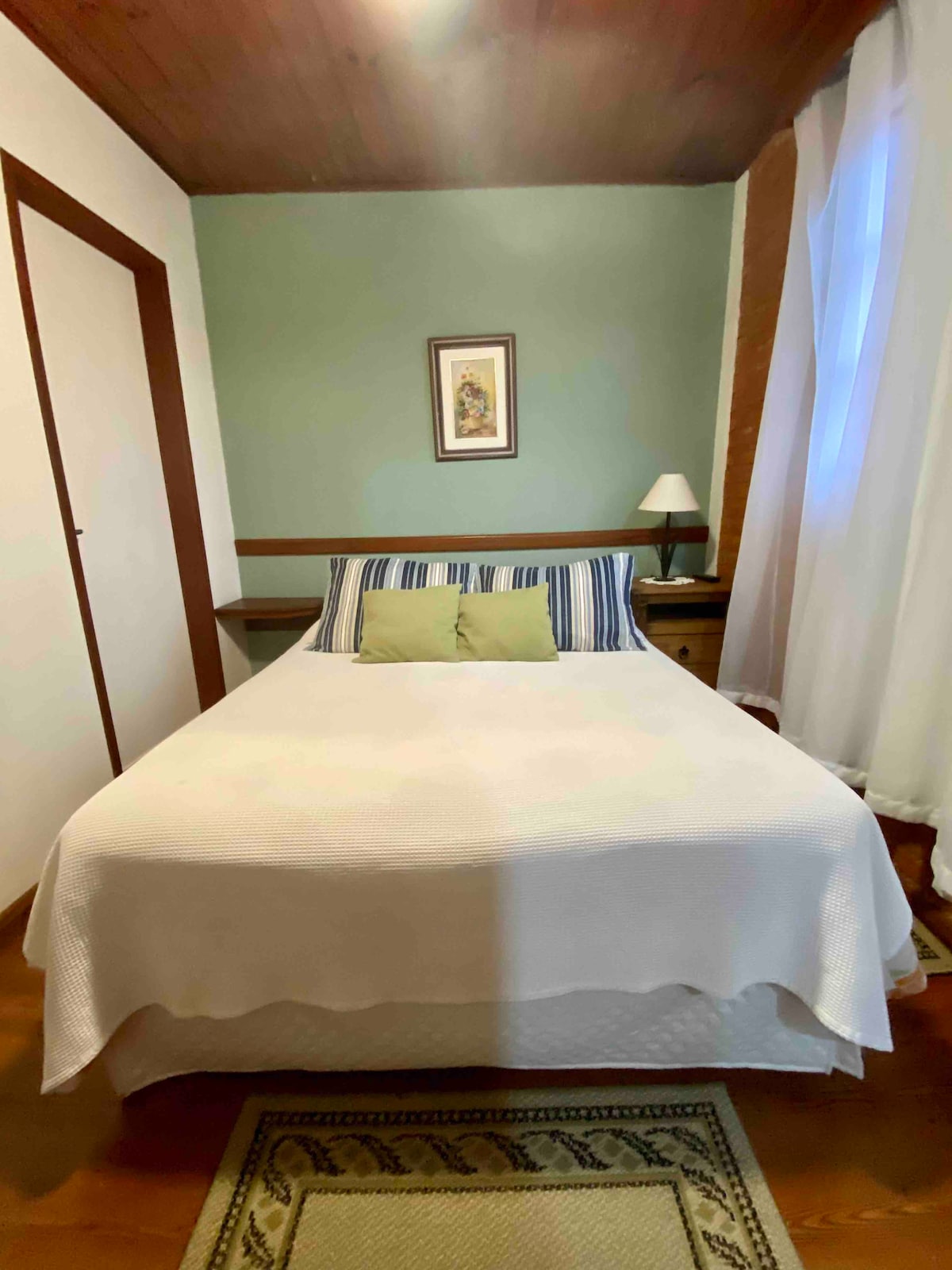 A cozy bedroom is presented with a spacious bed dressed in a light-colored coverlet. Paired striped pillows rest against a headboard. A bedside lamp and small table complement the room, while sheer curtains allow natural light to filter in, enhancing the relaxing ambiance.