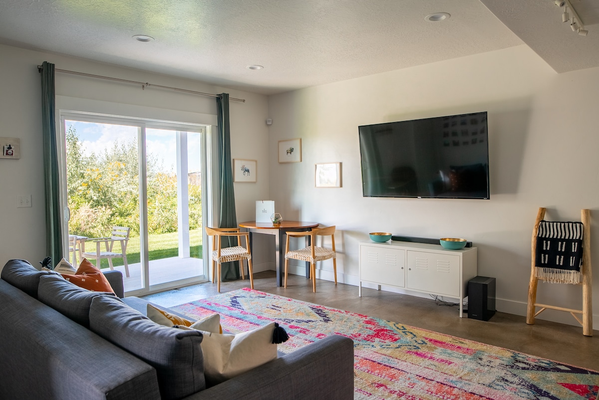 The main living area features a grey sofa facing a mounted television. A colorful area rug adds warmth to the space, complemented by two wooden tables and chairs. A sliding glass door opens to a grassy outdoor area, framed by light curtains.