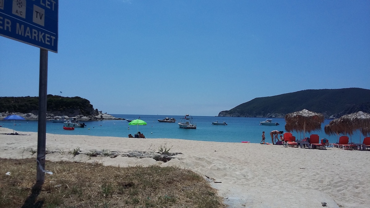 A sandy beach scene features colorful umbrellas and lounge chairs arranged along the shore. Calm turquoise waters reflect the sky, with boats anchored in the distance. Lush green hills frame the beach, creating a serene backdrop ideal for relaxation.