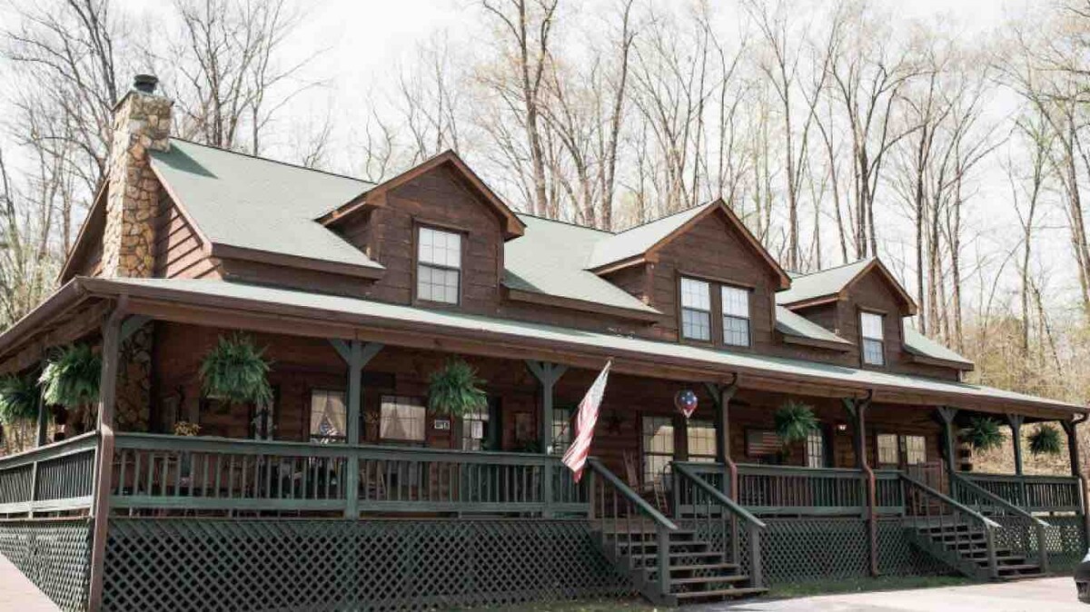 The exterior of a rustic log cabin is presented, featuring a spacious porch with a dark green railing. Large windows lined with potted ferns offer a welcoming facade. The building is nestled among trees, contributing to a serene outdoor setting.
