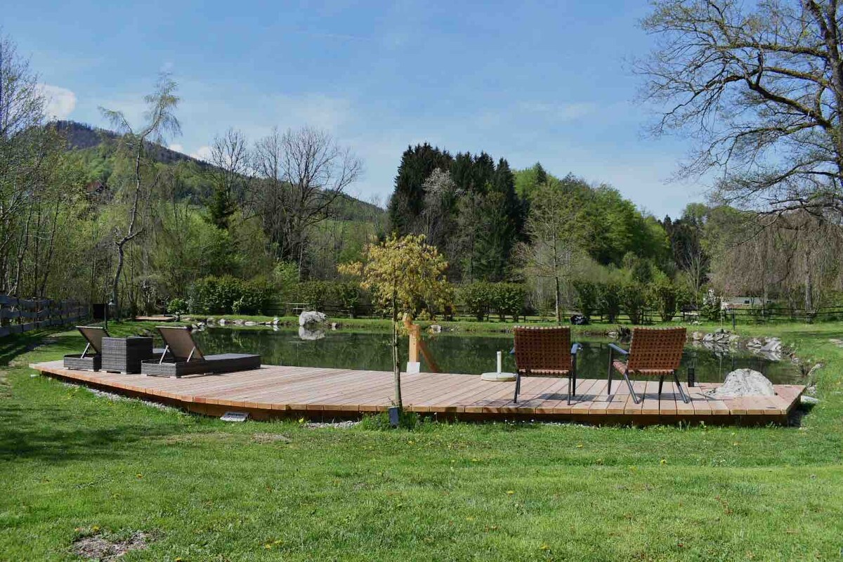 A spacious wooden deck overlooks a serene natural swimming pond, surrounded by lush greenery and trees. Two comfortable lounge chairs are positioned to enjoy the tranquil view, while a small table creates a cozy seating area. The vibrant landscape offers a peaceful ambiance.