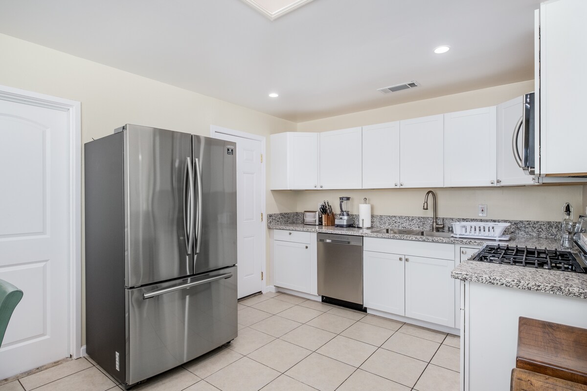 The kitchen features modern stainless steel appliances, including a large refrigerator and dishwasher. White cabinetry offers ample storage, complemented by a granite countertop. The space is well-lit with recessed lighting and tiled flooring, creating a functional cooking environment.