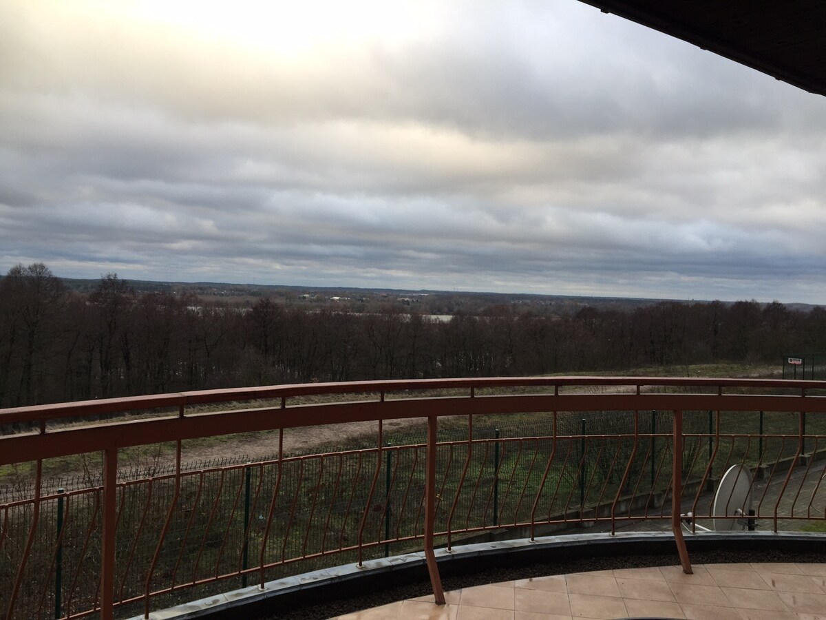 An outdoor terrace expands from the living area, featuring a curved railing. The view overlooks a wide landscape of trees and rolling hills, leading to a distant horizon with cloud coverage. The natural surroundings offer a serene backdrop for relaxation.