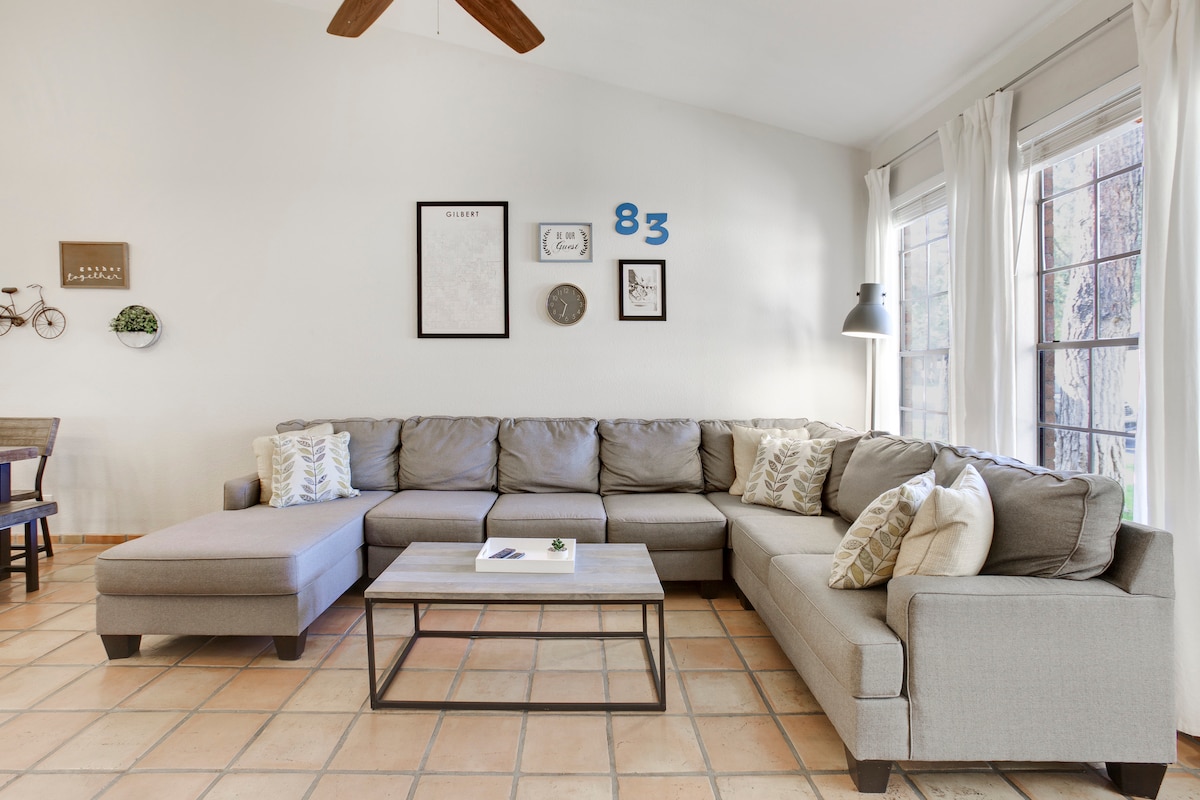A spacious living area features a large sectional sofa adorned with decorative pillows. A coffee table sits at the center, complemented by framed artwork on the walls. Natural light enters through tall windows, creating a bright and inviting space.