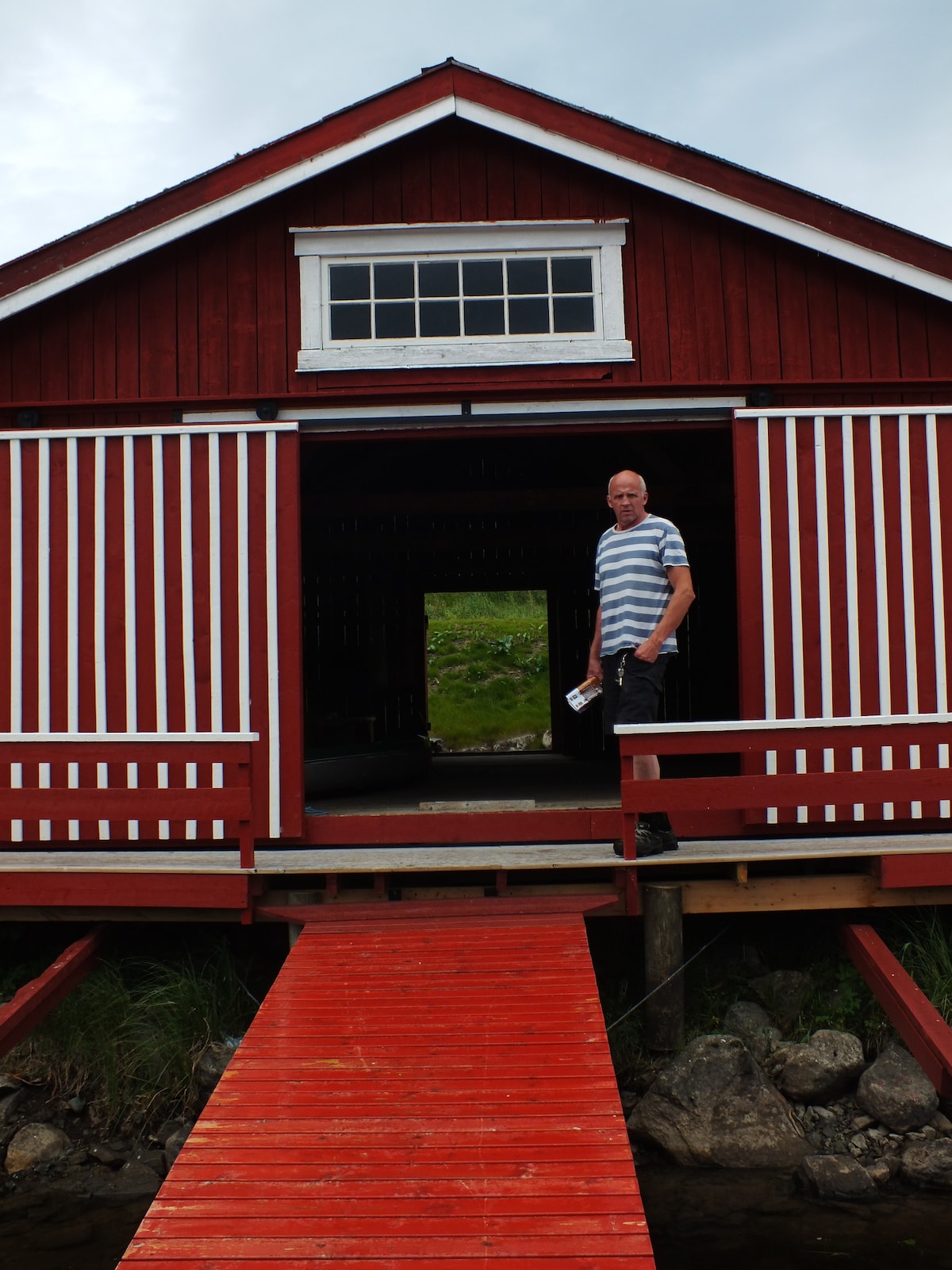 A red wooden boathouse features prominent white stripes, with a wide entrance revealing a clear view of the interior. A wooden walkway leads from the entrance toward the water, framed by grass and rocks, indicating easy access to outdoor activities.