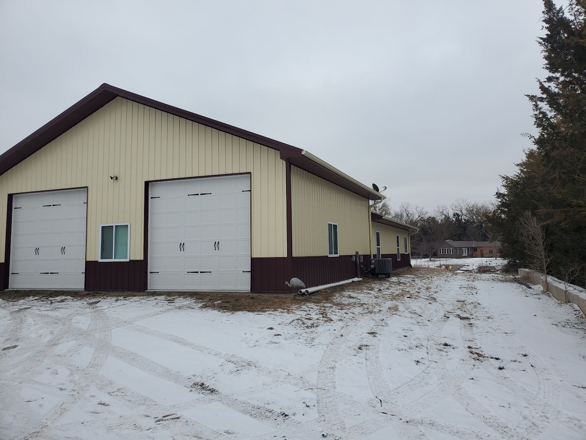A large, two-toned building is shown, featuring metal siding with a light-colored upper section and darker lower trim. Two overhead garage doors are visible on the front, along with windows on either side. The surrounding area is clear, with patches of snow covering the ground.