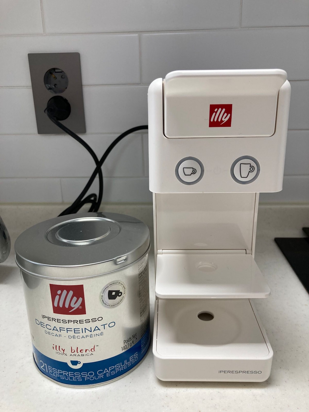 A countertop is displayed, featuring an Illy espresso machine beside a canister of decaffeinated coffee capsules. The machine is white, with buttons for coffee preparation, and the canister is metallic with a blue label. The area is well-lit and organized.