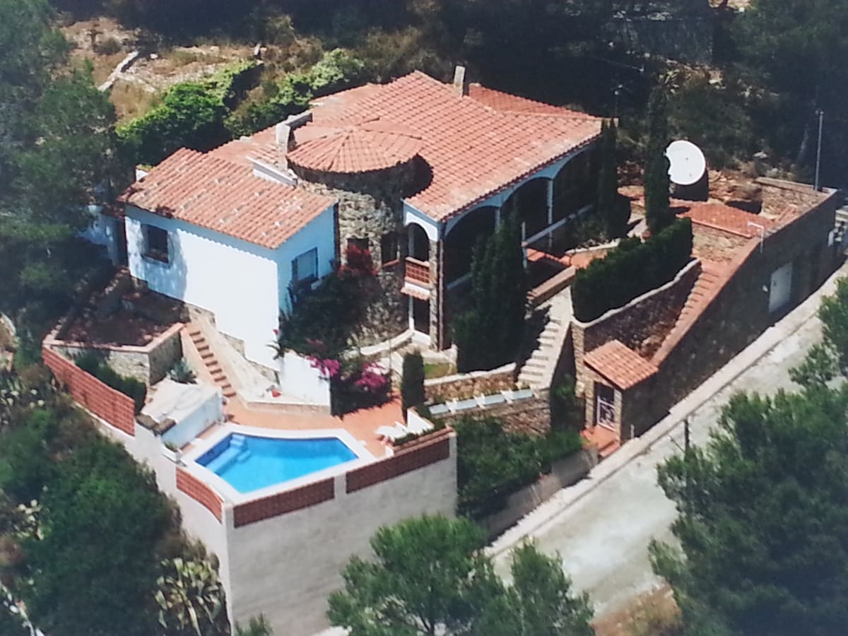 The property is presented from an aerial view, showcasing its unique architecture. The house features a circular roof element and terracotta tiles. Surrounded by greenery, a private pool can be seen adjacent to the house, accessible via landscaped terraces.