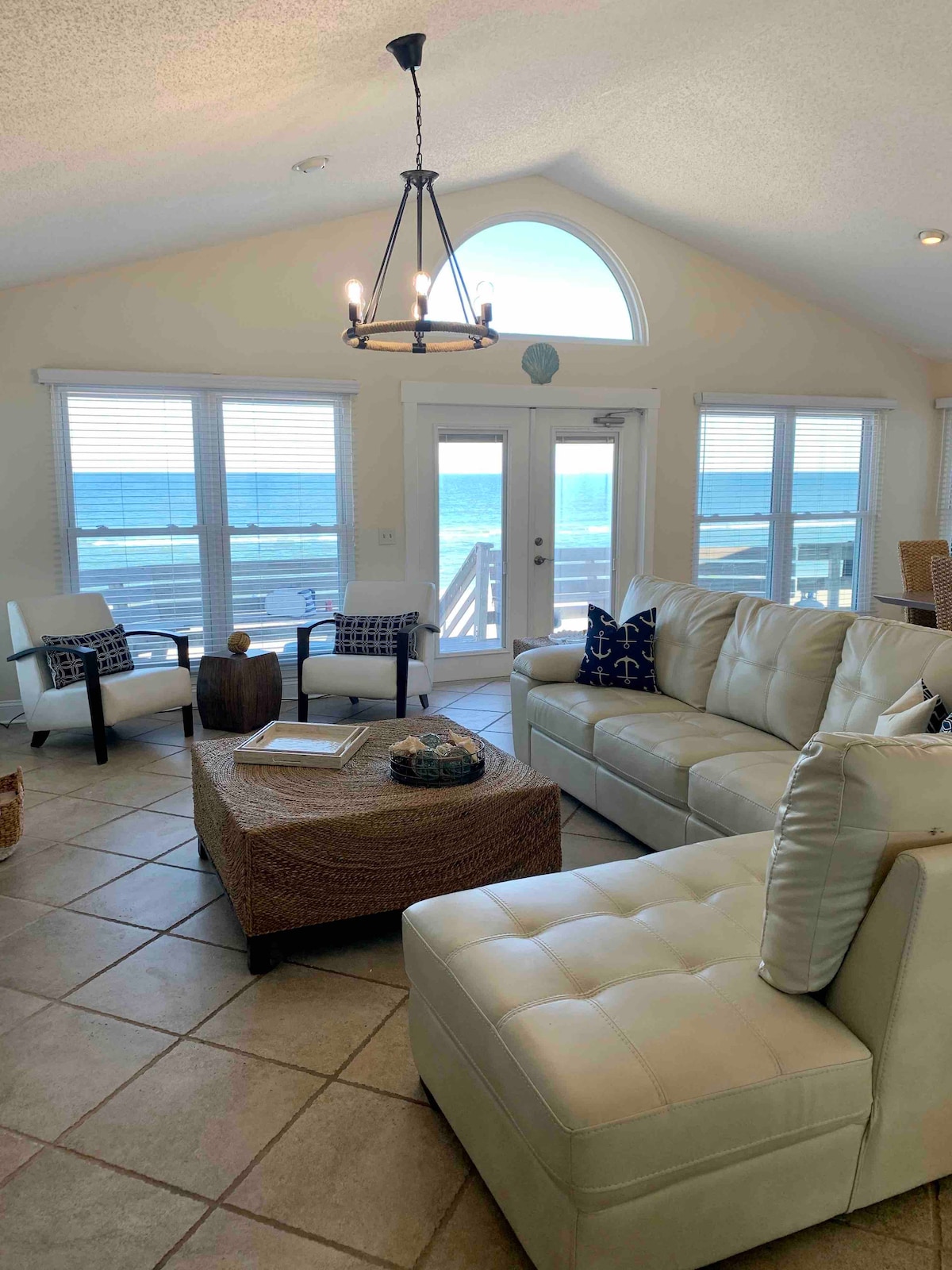 A bright living area features large windows that provide ocean views. A comfortable sectional sofa is accompanied by a woven ottoman and chairs, creating an inviting space. Natural light fills the room, highlighting the tile flooring and simple decor elements.