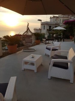 A spacious terrace features comfortable seating arrangements, including a hanging chair, white wicker chairs, and a small table. Potted plants add a touch of greenery. The scene is illuminated by the warm glow of the sunset over the sea, creating a tranquil ambiance.