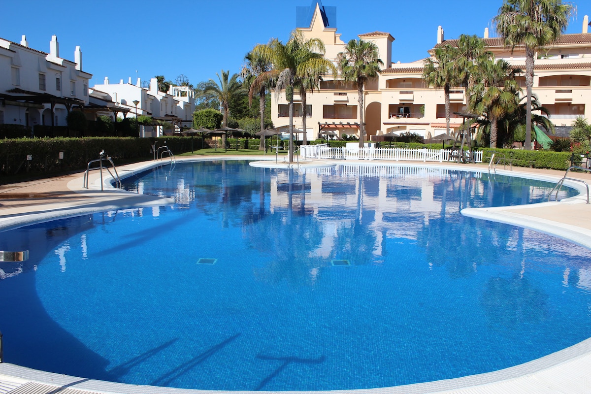 A large swimming pool is centrally located within a well-maintained garden area. Sunlight reflects off the crystal-clear water, while palm trees are positioned around the pool's edge. Lounge chairs and shaded areas are visible, enhancing the relaxed atmosphere of the surrounding space.