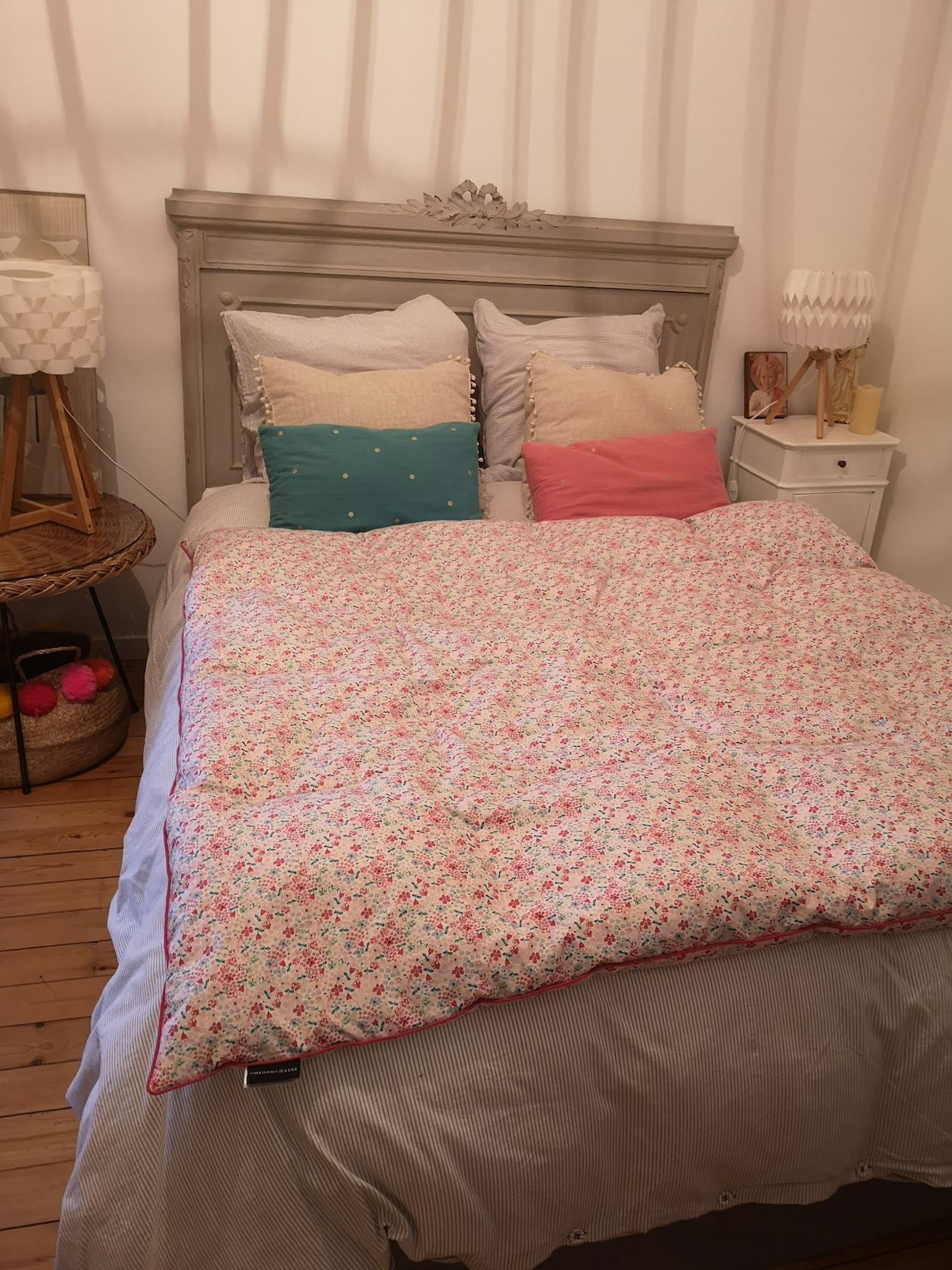 A spacious bed is dressed with a floral patterned comforter and several decorative pillows in various colors. A wicker basket and a lamp are positioned beside the bed on a small table, while a nightstand holds a minimalist lamp and decor, enhancing the cozy atmosphere.
