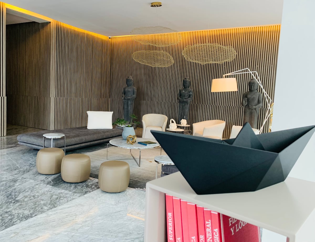 A modern lobby area is depicted, featuring a minimalist design with wooden accents and elegant lighting. Comfortable seating is arranged around a glass table, accompanied by decorative sculptures and a striking black artwork resembling a boat. Soft, neutral tones create a serene ambiance.
