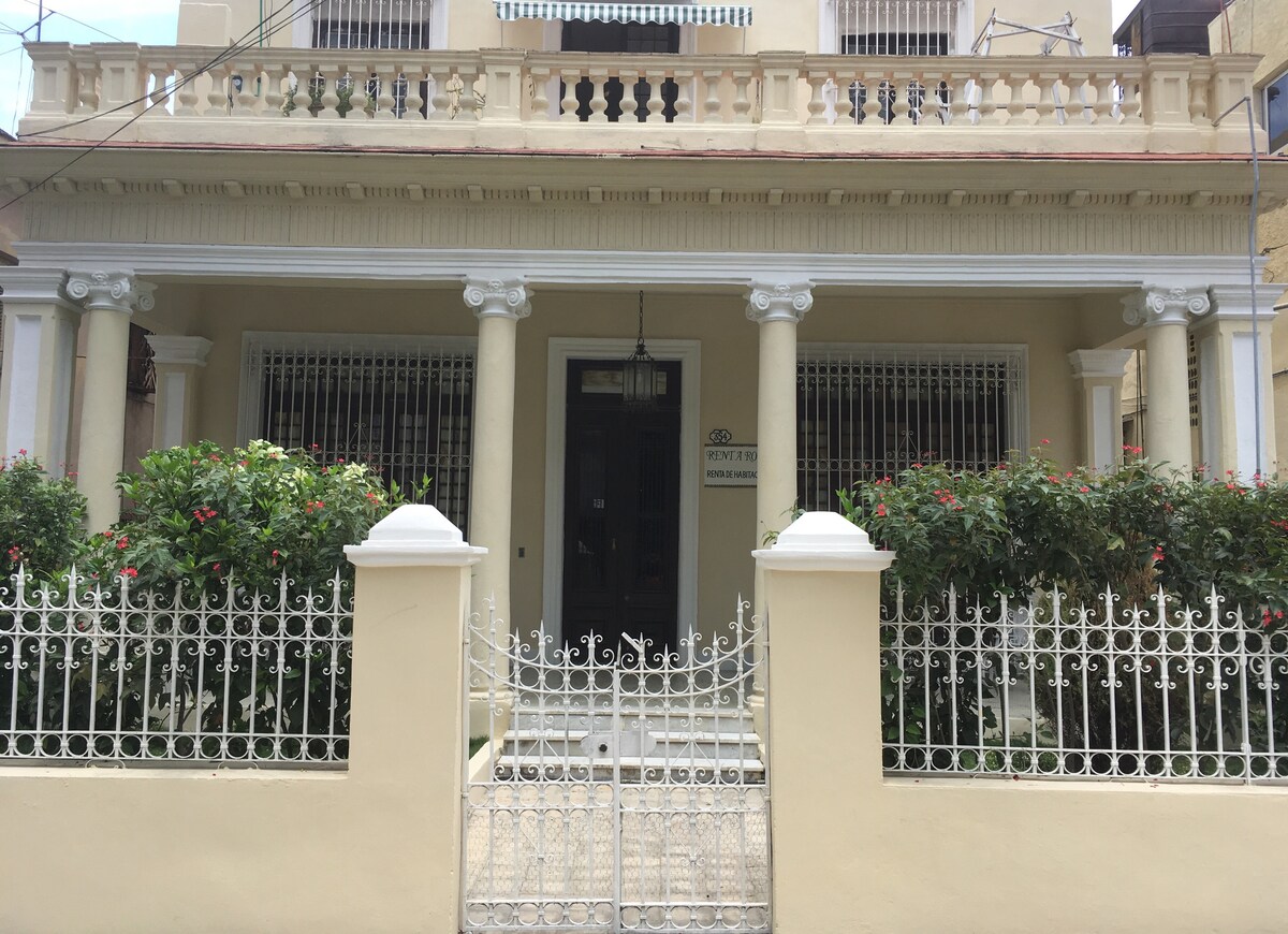The exterior of a colonial-style boutique hostal is presented, featuring a light-colored façade with decorative columns and a wrought-iron gate. Lush greenery flanks the entrance, and a balcony is visible above the front porch, contributing to the architectural charm.