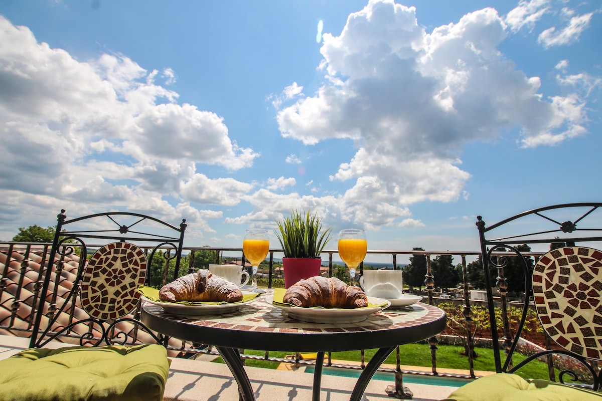 An outdoor balcony table is set with two croissants and glasses of orange juice. Two wrought iron chairs line the sides. A small green plant sits in a pot at the center, with a clear blue sky and fluffy clouds visible in the background.