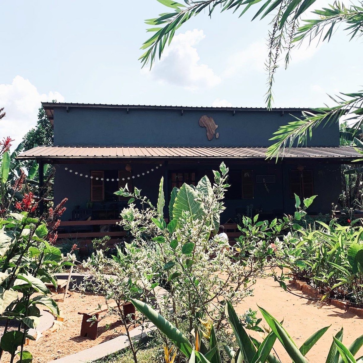 A modern house is set amidst lush greenery, showcasing a vibrant garden filled with various plants and flowers. The structure features a covered porch, with seating arranged to invite relaxation. Natural sunlight illuminates the exterior, enhancing the tranquil outdoor environment.