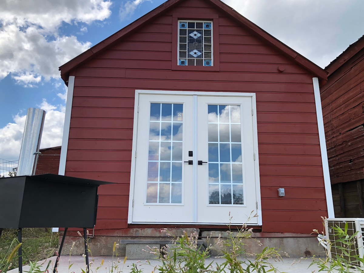The charming red cottage features a gabled roof and large double French doors framed by decorative windows. The sky, dotted with fluffy clouds, sets a serene backdrop. A minimalist grill is positioned nearby, enhancing the outdoor gathering space.