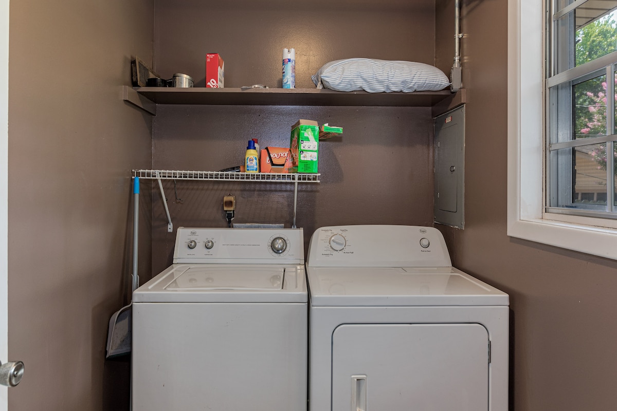 A laundry area is equipped with a washer and dryer, positioned side by side against a brown wall. Above the appliances, shelving holds various cleaning supplies and personal items, while a small window allows natural light to enter the space.