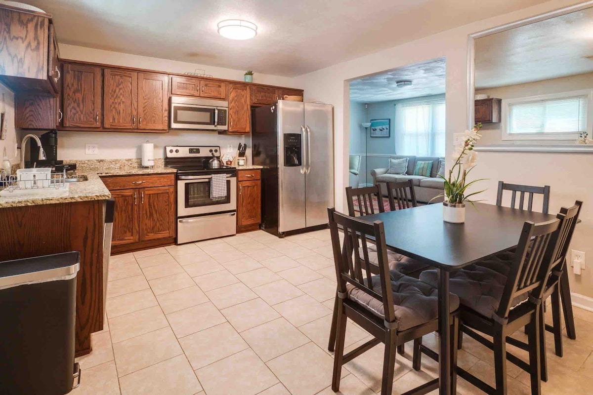 The kitchen showcases dark wood cabinetry with granite countertops, complemented by stainless steel appliances. A dining table with four chairs sits nearby. Natural light enters through a window, illuminating the tiled floor and providing a welcoming atmosphere.