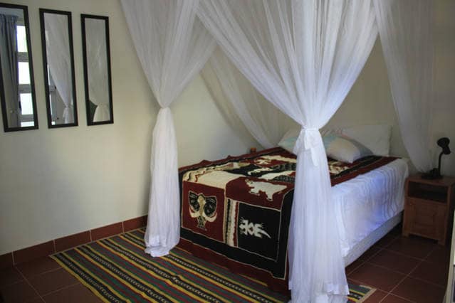 A spacious bed is draped with a colorful blanket, surrounded by sheer white mosquito netting. The room features three wall-mounted mirrors and a warm-toned area rug beneath the bed, with soft lighting from a nearby lamp enhancing the serene ambiance.