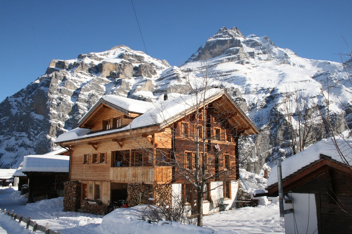 A traditional chalet, constructed with wooden materials, is situated in a snowy landscape. The structure features multiple balconies and stone accents, surrounded by snow-covered ground. Towering mountains form a striking backdrop, creating a serene winter atmosphere.