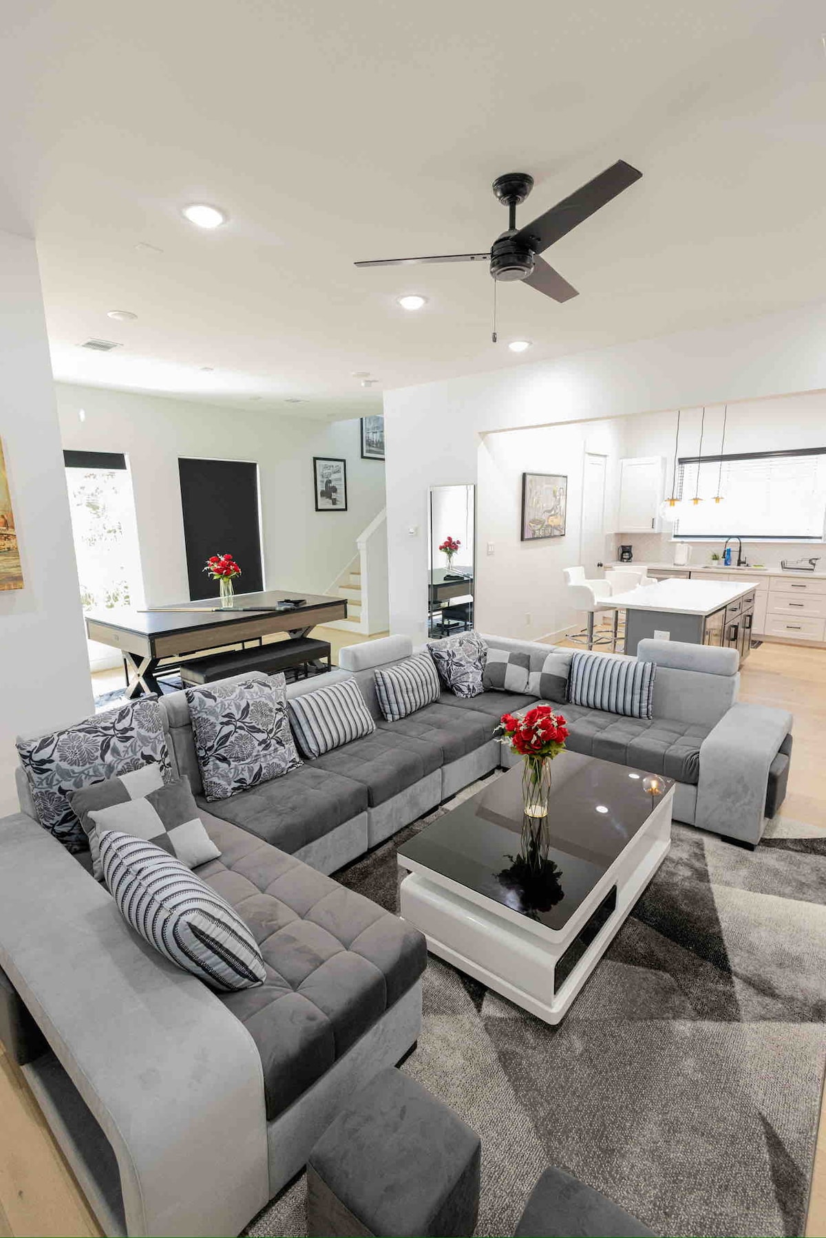 A modern living area features a spacious sectional sofa adorned with accent cushions and a glass coffee table. The open-concept design highlights natural light, with a dining area and fully equipped kitchen visible in the background, complemented by subtle artwork on the walls.