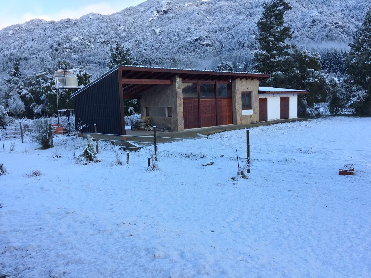 A spacious house is set against a snowy landscape, with a combination of stone and modern materials. Large glass doors are visible, offering a view of the surrounding snow-covered grounds. The property features a covered area on one side, along with an open grassy space.