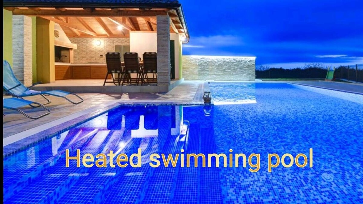 A heated swimming pool reflects the evening sky, surrounded by a spacious terrace. Nearby, a summer kitchen with a dining area is visible under the shelter of a wooden roof, offering a comfortable gathering space.