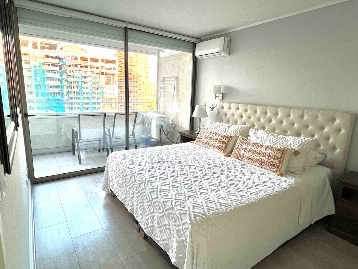 A bright bedroom is shown with a spacious bed featuring a tufted headboard. Large windows offer views of the city skyline. A small table and chairs are visible on the balcony. Light-colored walls and wooden flooring create a calm and modern feel.