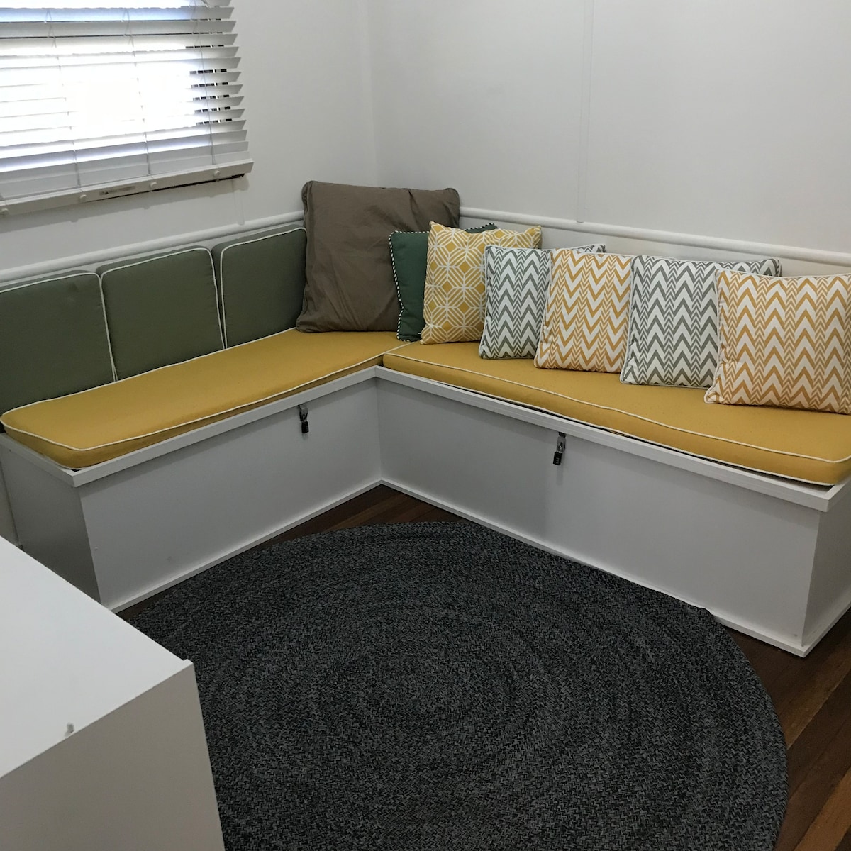 A corner seating area features a yellow and green cushioned bench with several textured throw pillows in various patterns. The space is complemented by a round, braided rug beneath, with sunlight filtering through the nearby window adorned with white blinds.