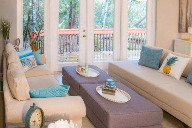 A spacious living area is highlighted by modern, light cream sofas adorned with decorative pillows in soft blues and greens. A large, rectangular ottoman centers the room, displaying various decorative items. Glass doors open to a wooden deck, offering a view of the lush outdoor landscape.