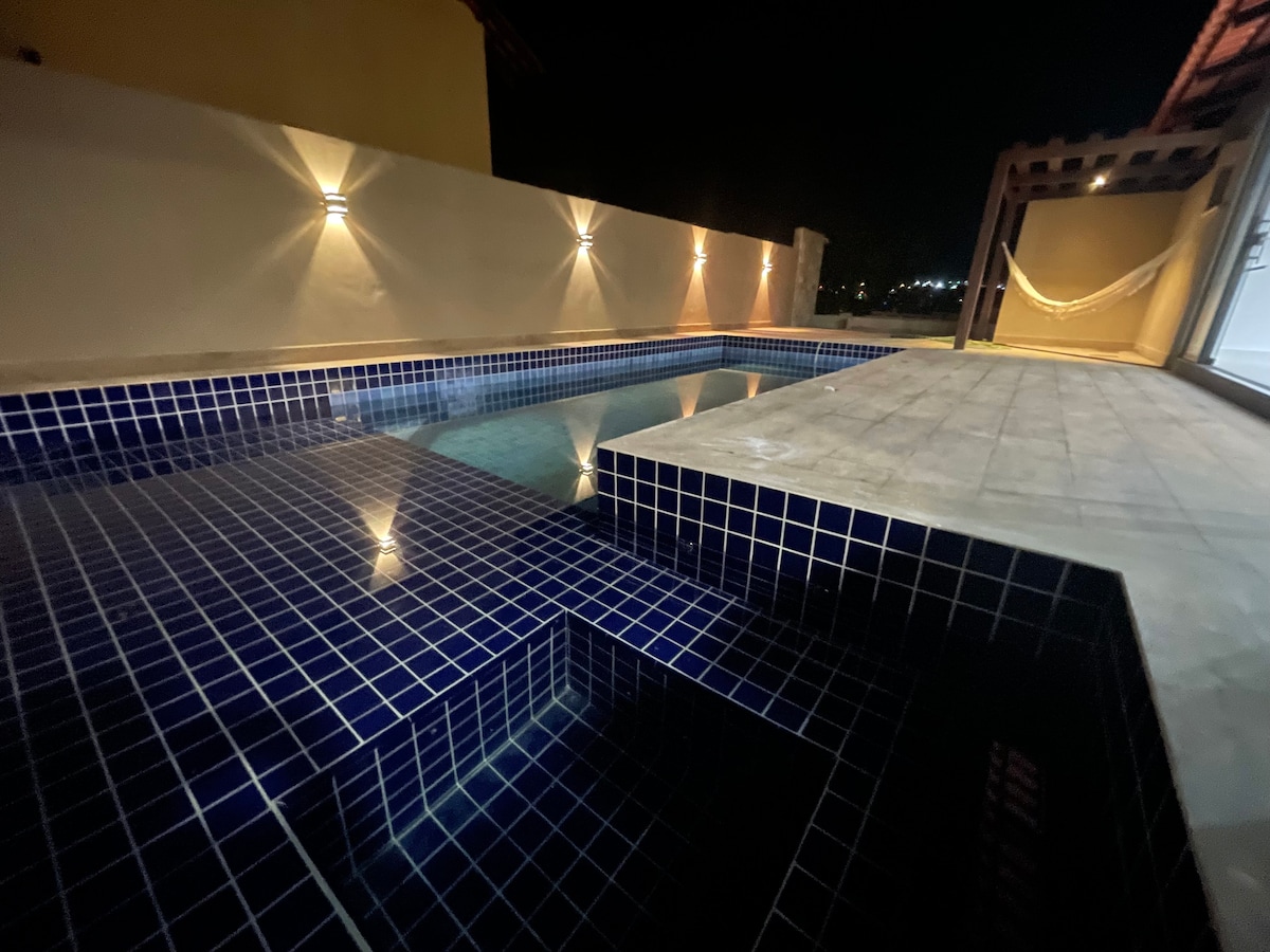 A serene pool area is illuminated at night, featuring a vibrant blue tile surface. The heated pool includes a separate section for hydromassage, with soothing water reflections. The surrounding deck is spacious, complemented by ambient lighting along the walls.